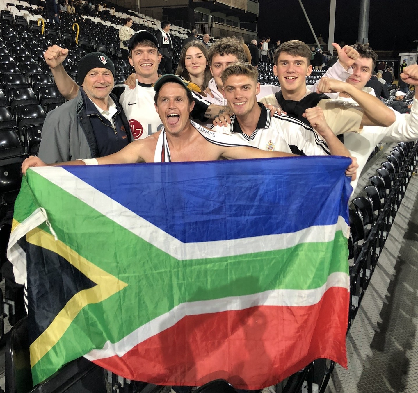 Chairman of the Fulham Supporters Club in SA, James Preston, holding up an SA flag at Fulham's stadium, Craven Cottage along with other fans.